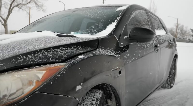 A dark-colored car, heavily coated in fresh snow, sits parked on a snowy day