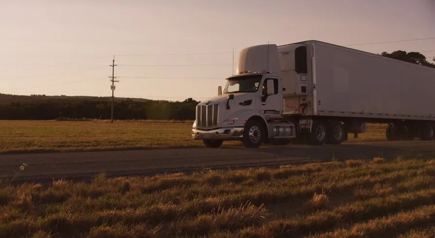 A large, light-gray semi-truck travels down a rural road at sunset