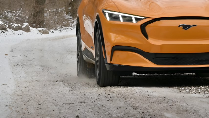 A vibrant orange Ford Mustang Mach-E navigates a snowy, gravel road