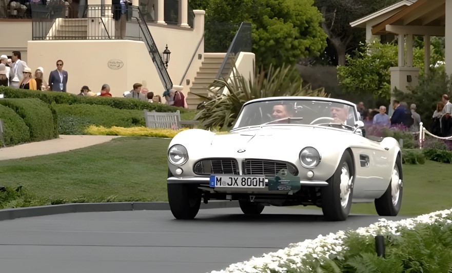 A classic car drives down a road while onlookers watch from the sidelines