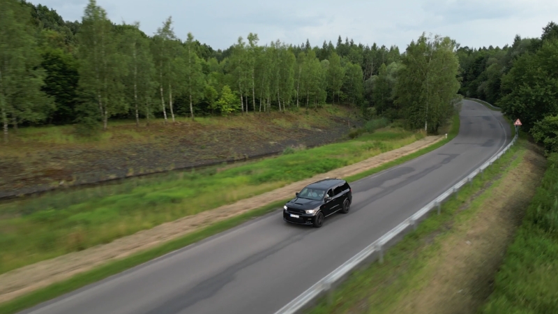 A black car drives along a quiet, curving road surrounded by trees in rural Europe