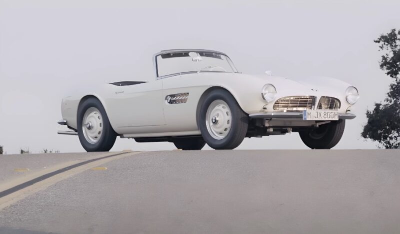 A classic white BMW 507 Roadster car driving along a scenic road under clear blue skies