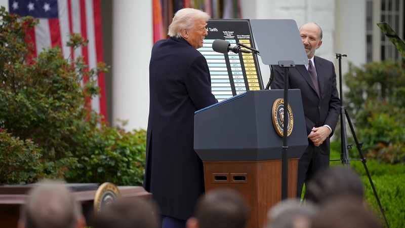 U.S. President speaking at a press event with a tariff chart in front of an audience