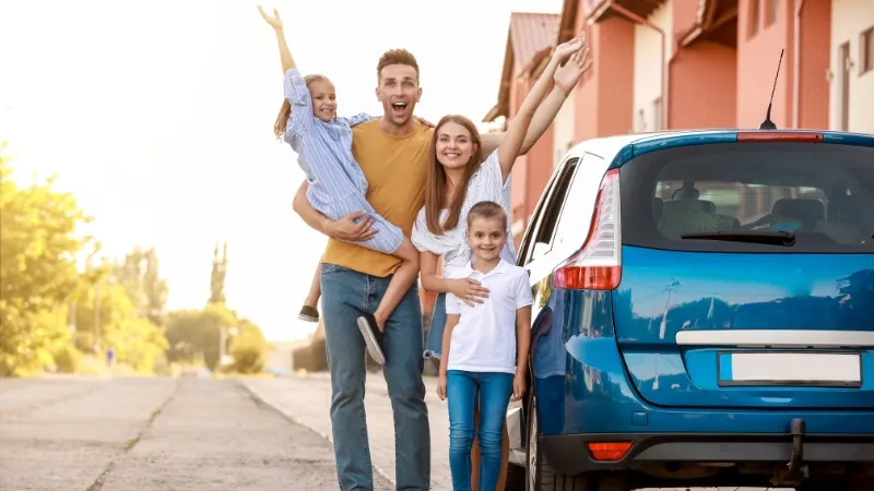 A happy family stands next to a blue car, celebrating one of the safest family cars