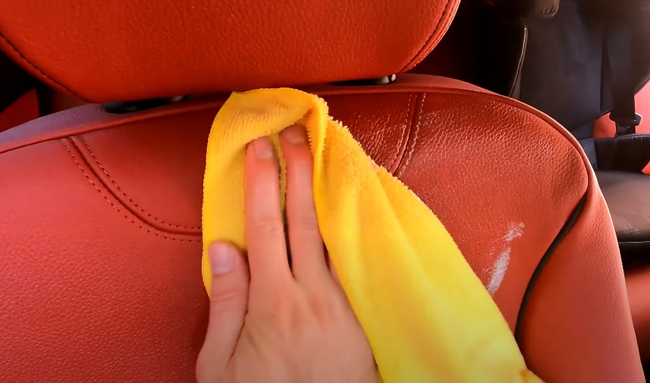 A hand cleans a red leather car seat with a yellow microfiber cloth