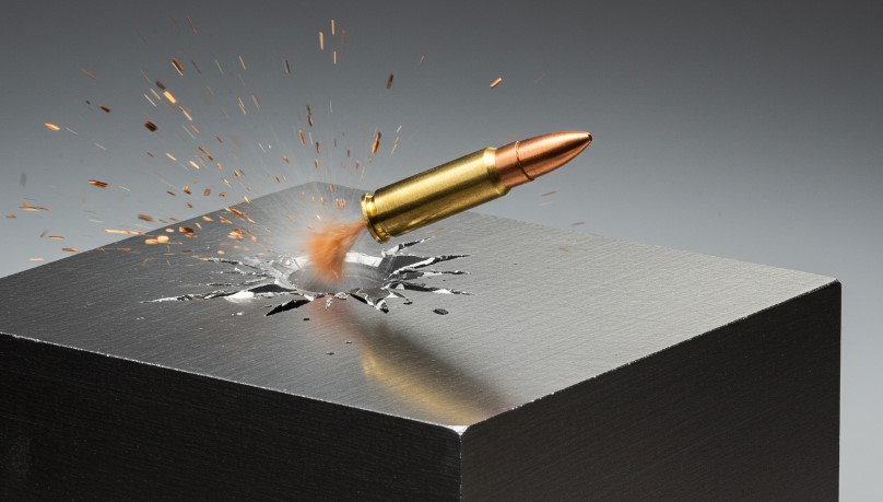 A bullet pierces a metal block, creating a dramatic impact crater