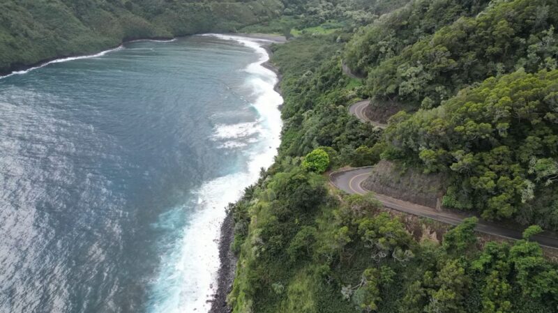 Aerial view of the winding Road to Hana along Maui’s lush coastline with ocean waves crashing below.