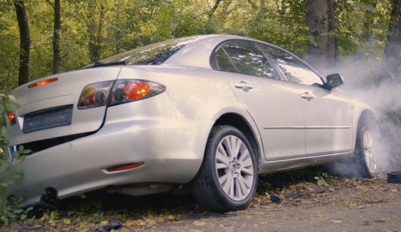 A silver sedan, with visible damage, sits in a wooded area