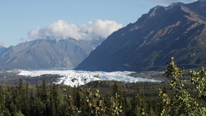 Snow-covered glacier beneath towering mountain peaks along the Richardson Highway in Alaska, framed by evergreen forest.
