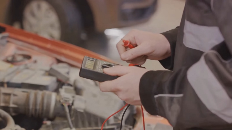 A mechanic holds a multimeter to test a car battery and charging system after an alternator replacement