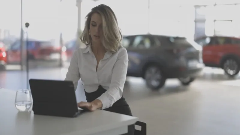 A woman works on a tablet inside a dealership with cars displayed in the background