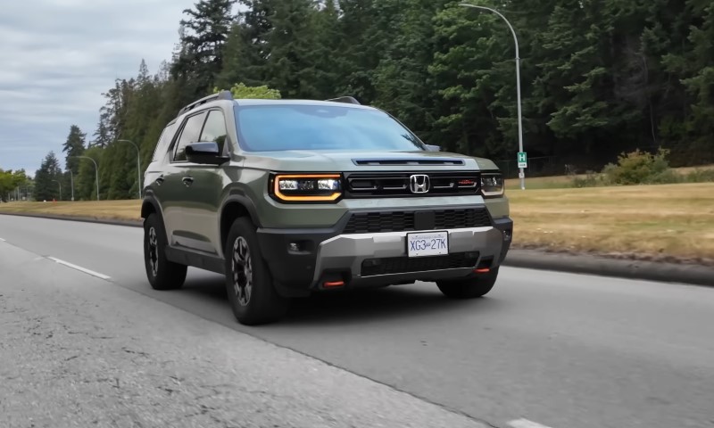 Green Honda Passport SUV driving on a highway bordered by lush trees, under an overcast sky