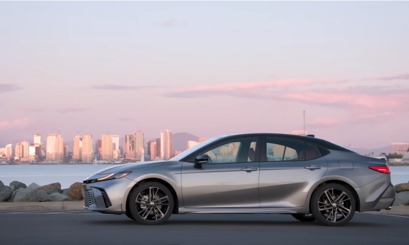 A sleek silver Toyota Camry sedan parked by a waterfront with a city skyline in the background under a pink-hued sky