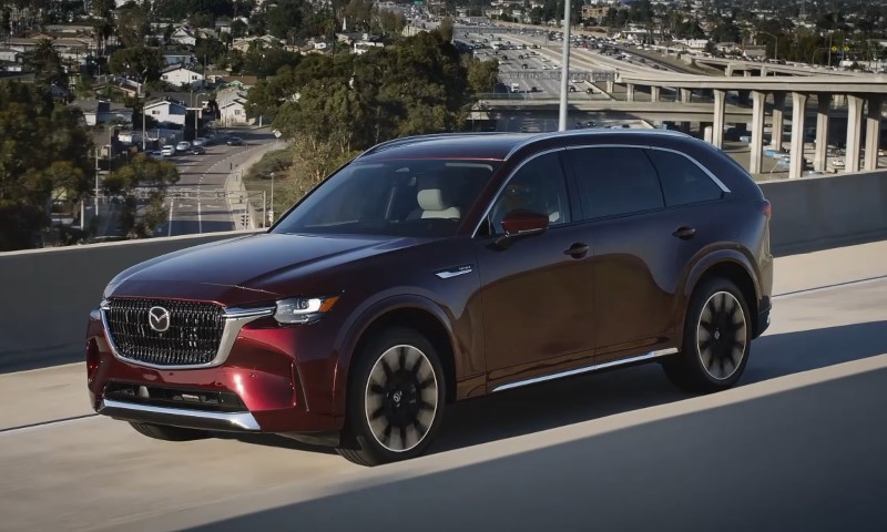 Red Mazda CX-90 SUV driving on a highway overpass in an urban setting, with a cityscape and greenery in the background